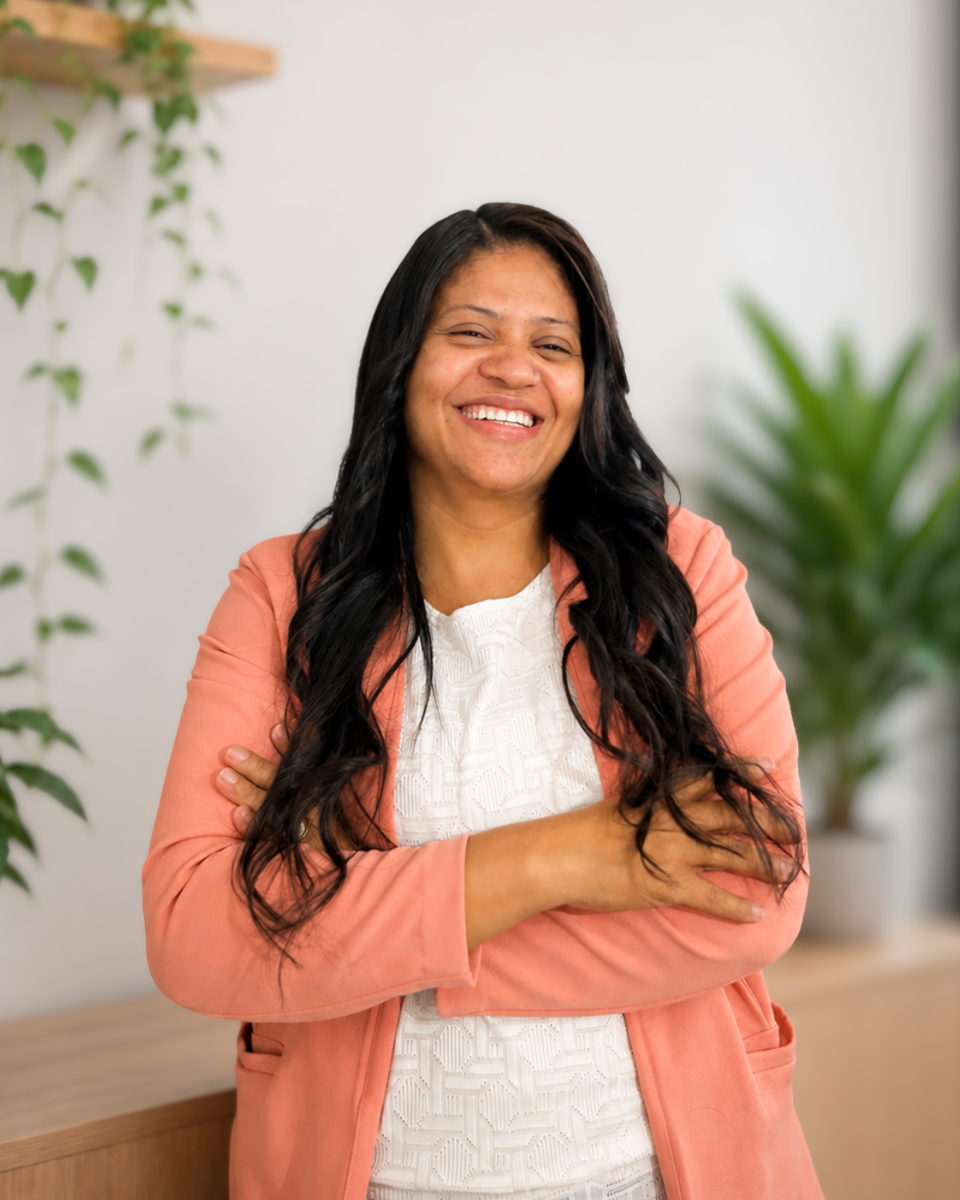 Cristiane Rosa, psicopedagoga, em pé e sorrindo, de blazer salmão sobre blusa branca, com plantas ao fundo.